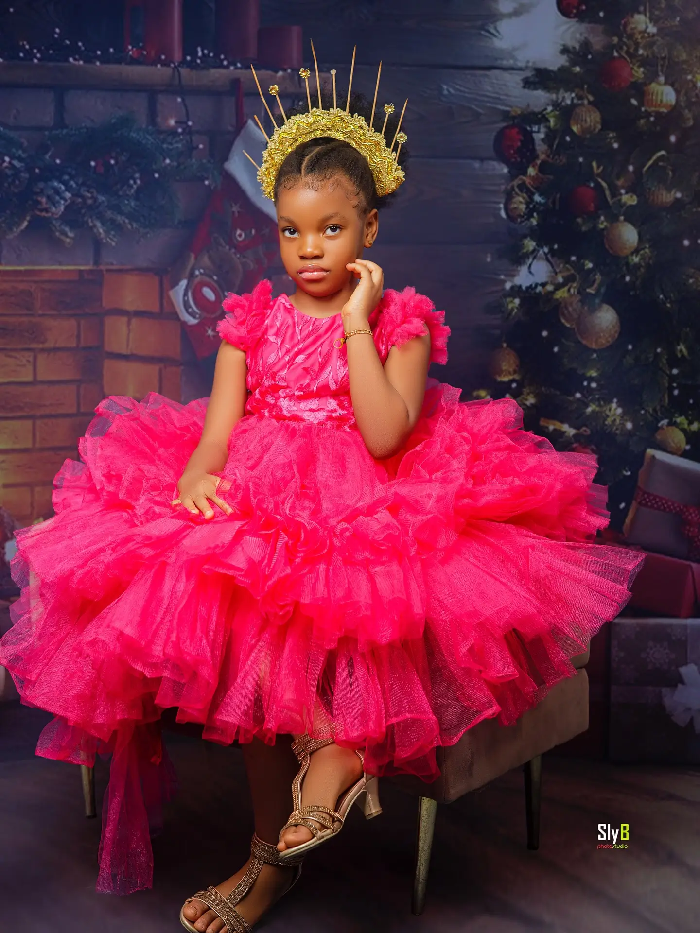 Girl in a pink tulle dress and gold crown seated in front of a Christmas themed backdrop.