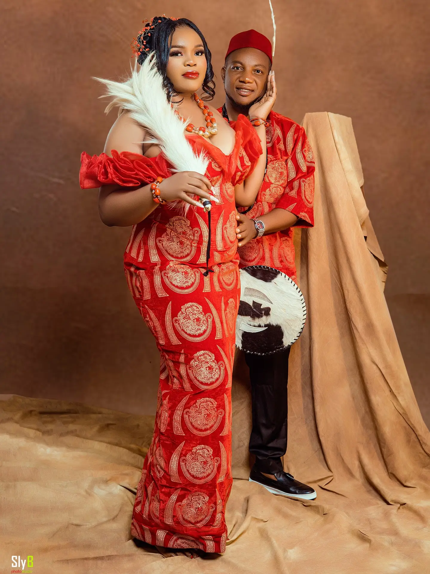 Couple in red traditional attire posing together with a feather fan and cow-skin drum.
