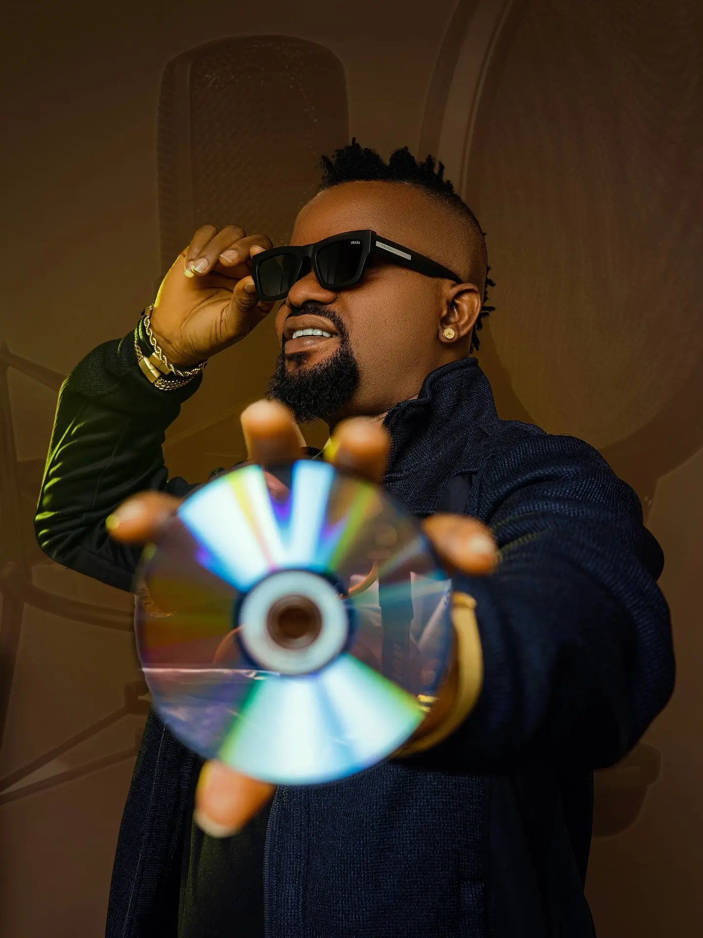 Man in a navy jacket and sunglasses holding a reflective CD toward the camera.