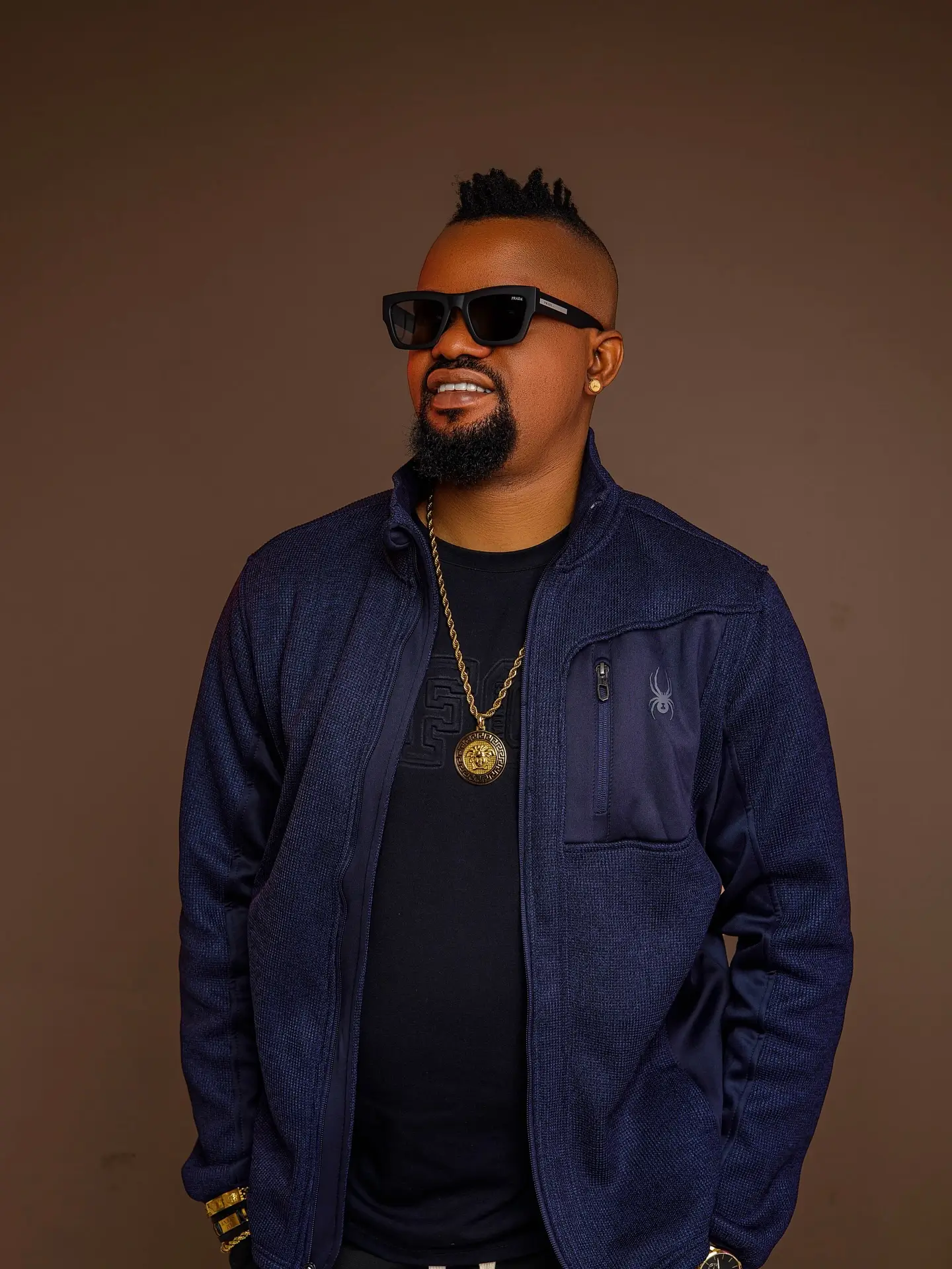 Man in a navy jacket and sunglasses standing against a brown backdrop.