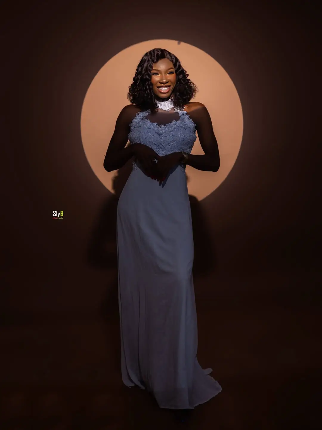 Woman in a blue gown with lace bodice smiling under a spotlight.