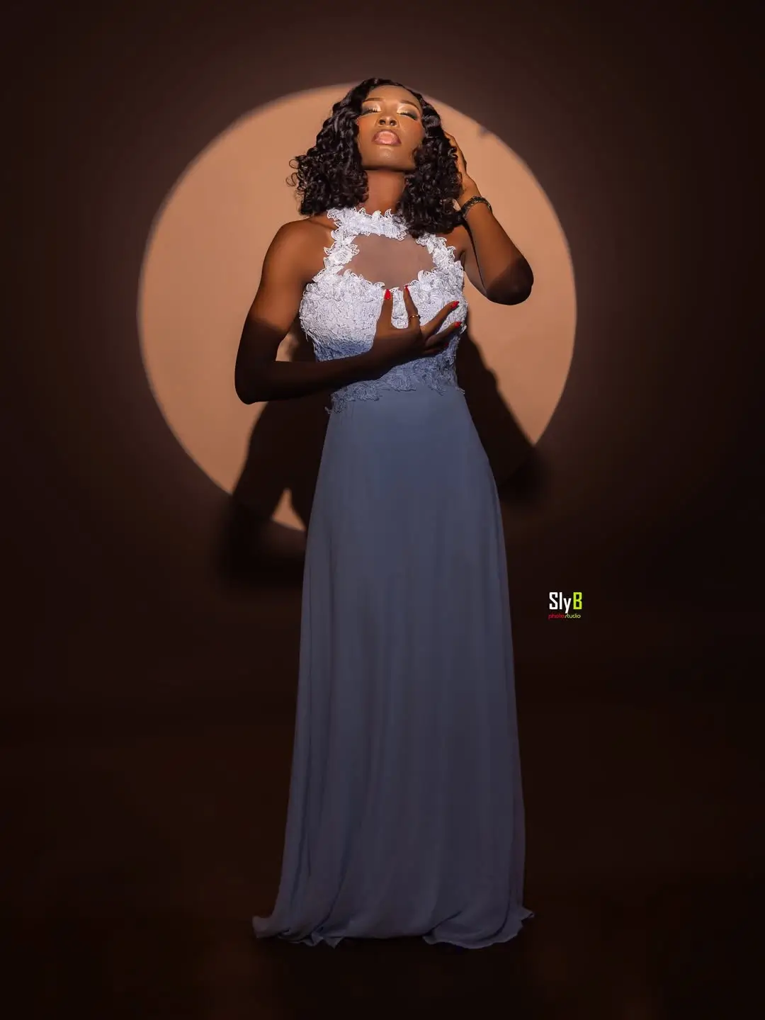 Woman in a blue gown with white lace detail standing in a circular spotlight.