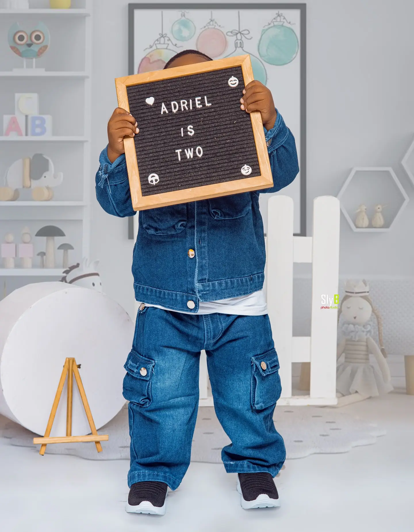 Toddler boy in a denim jacket holding a letter board that reads Adriel is two.