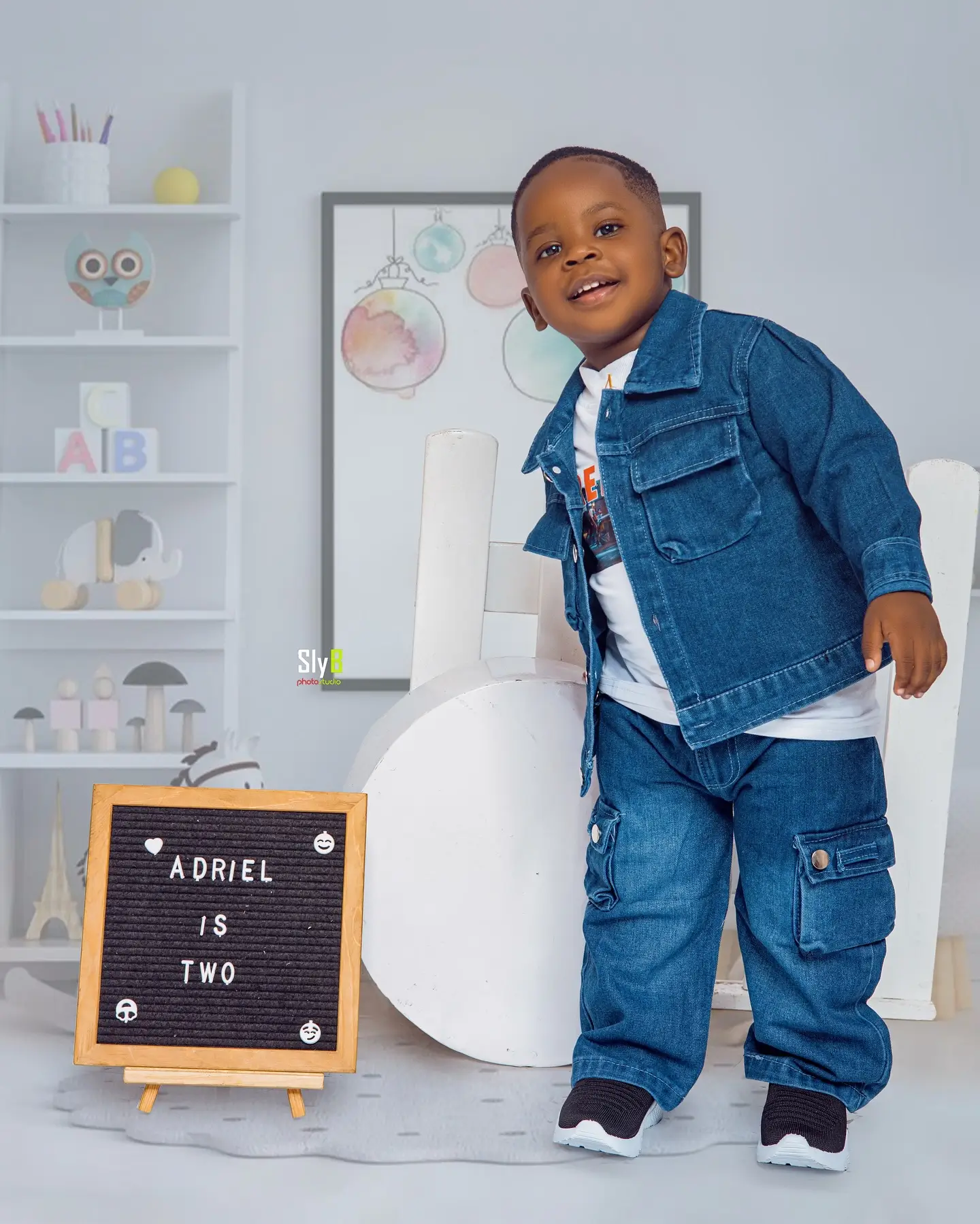 Toddler boy in a denim jacket and jeans smiling in a nursery-themed photo set.