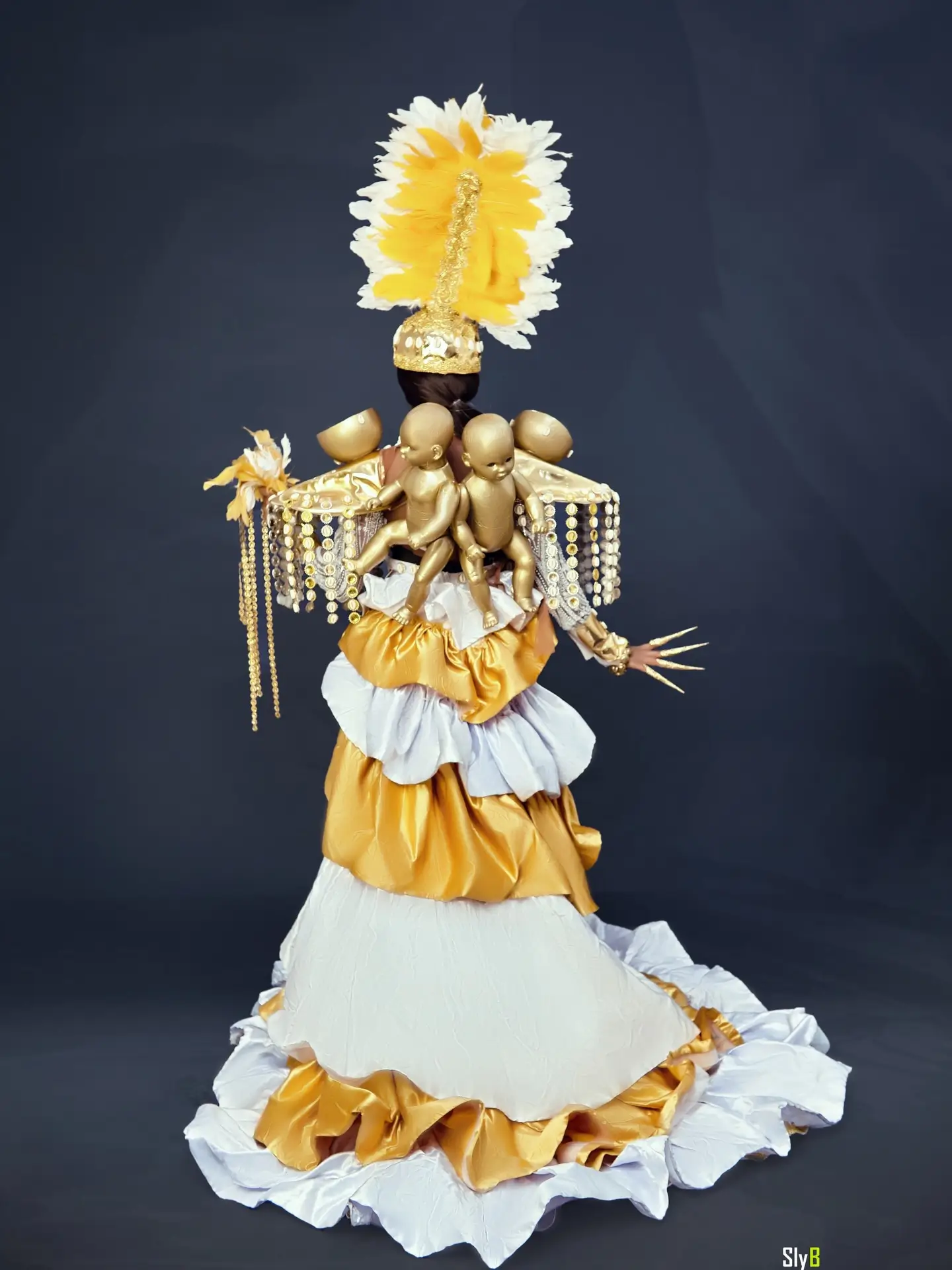 Back view of a woman in a gold and white costume with feather headdress and doll figures on her shoulders.