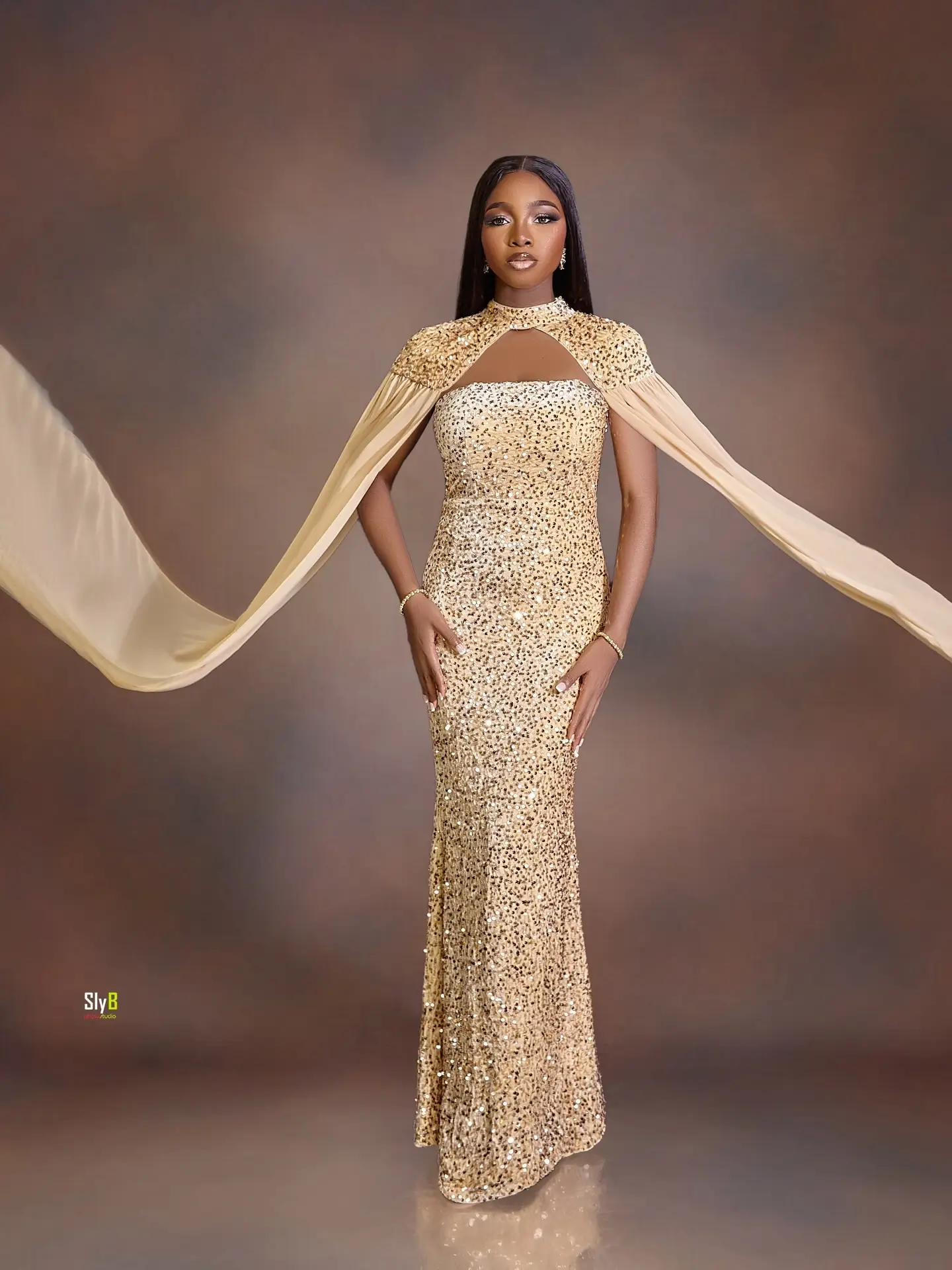 Woman in a gold sequin gown with flowing cape sleeves posing against a brown backdrop.