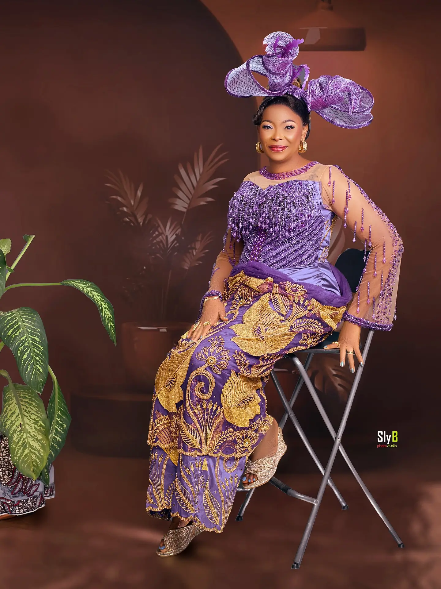 Woman in a purple and gold traditional outfit seated on a folding chair beside a plant.