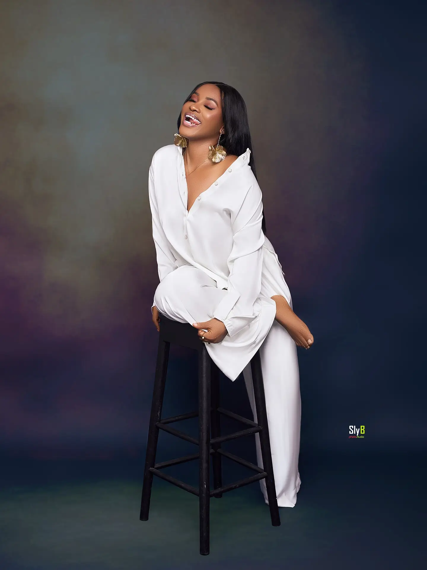 Woman in a white blouse and trousers smiling with her eyes closed on a stool.