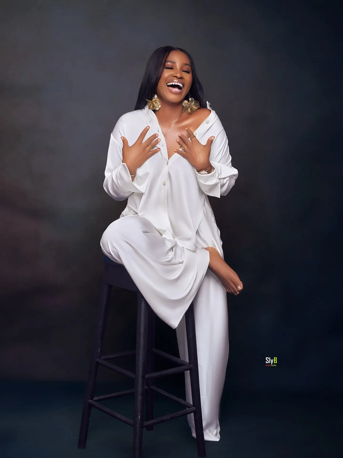 Woman in a white outfit laughing while seated on a black stool in a studio.