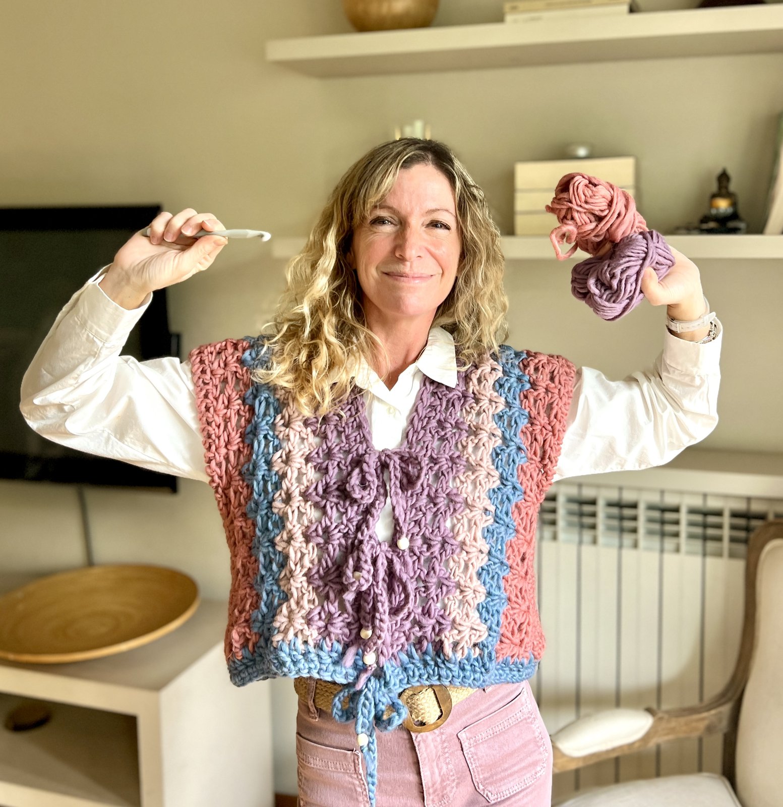 Trini sonriendo con un chaleco de crochet lila, con los brazos en alto, un ganchillo en una mano y ovillos en la otra