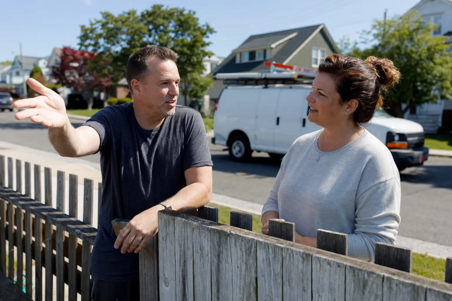 Two neighbors talking over a shared fence line or in a driveway, one gesturing toward their own house Two neighbors talking over a shared fence line or in a driveway, one gesturing toward their own house