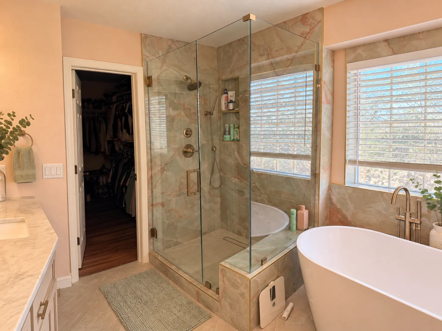A modern bathroom features a double vanity with light wood cabinets, gold fixtures, and a glass-enclosed shower, with a view into a bedroom beyond.