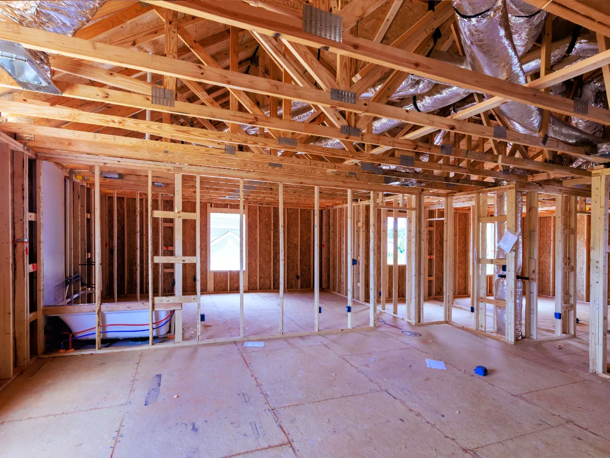 Interior view of a new house under construction, showing exposed wooden wall studs and roof trusses with insulation.