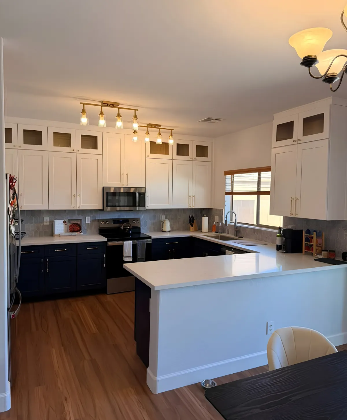 A modern kitchen with white upper cabinets, dark blue lower cabinets, white countertops, wood flooring, and gold light fixtures.
