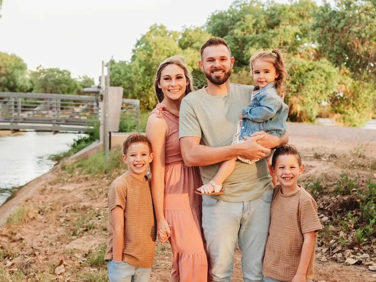 A smiling family of five, including parents and three children, poses outdoors by a canal with green trees and a bridge.