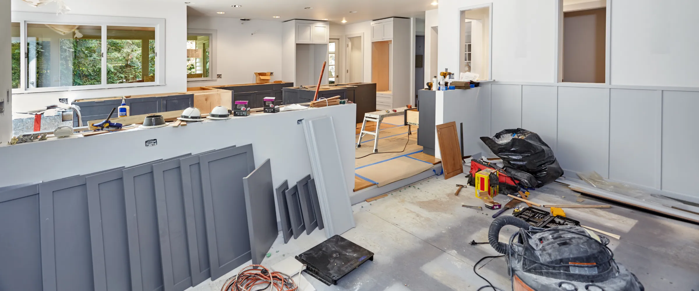 A wide shot of a kitchen under construction, featuring gray cabinets, various tools, and building materials scattered around.