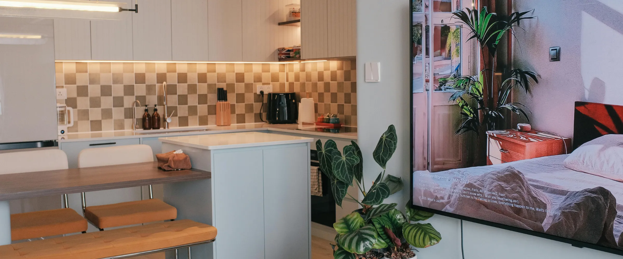A modern open-plan kitchen and dining area with light cabinets and checkered backsplash, adjacent to a TV displaying a bedroom scene and a large potted plant.