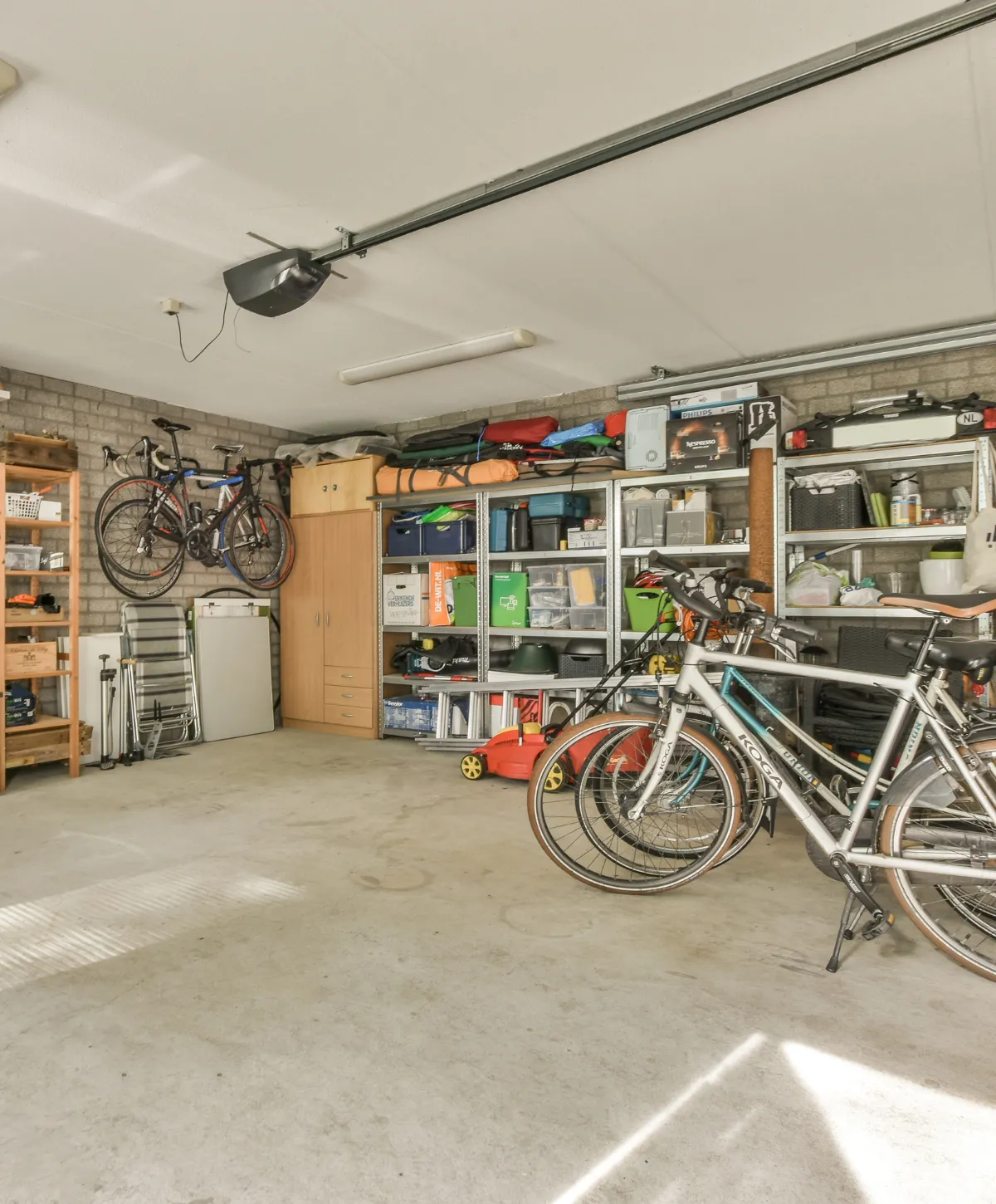 An organized garage features multiple bicycles, storage shelves with various items, and a concrete floor with sunlight patterns.
