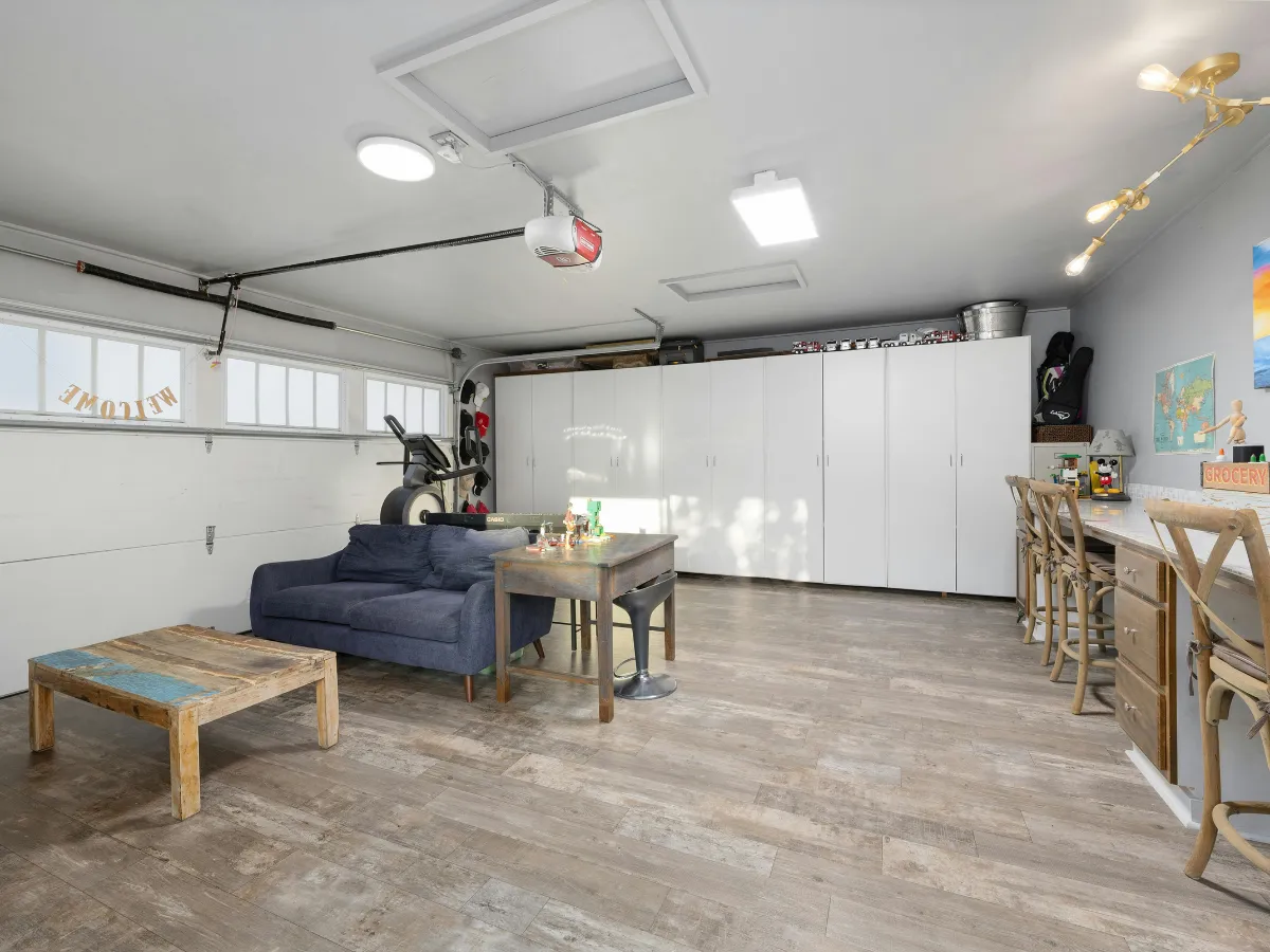 A brightly lit, converted garage featuring a blue sofa, bar area with stools, and extensive white storage cabinets.