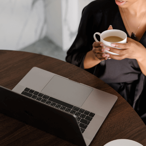 Woman of color holding a cup of coffee while looking at a laptop