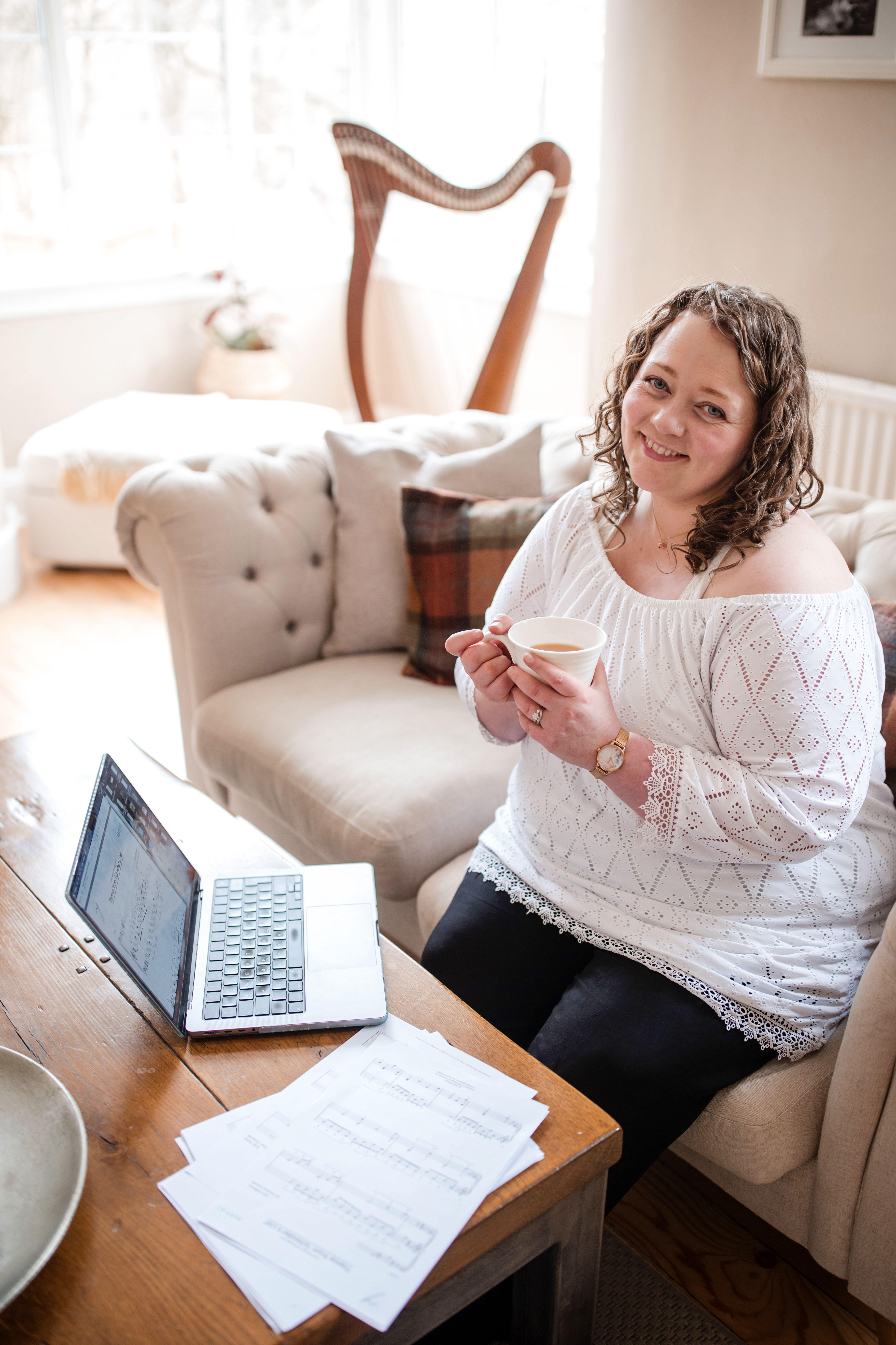 Heather sitting at her desk, cup of tea in hand, harp in the background and music sheets on the table