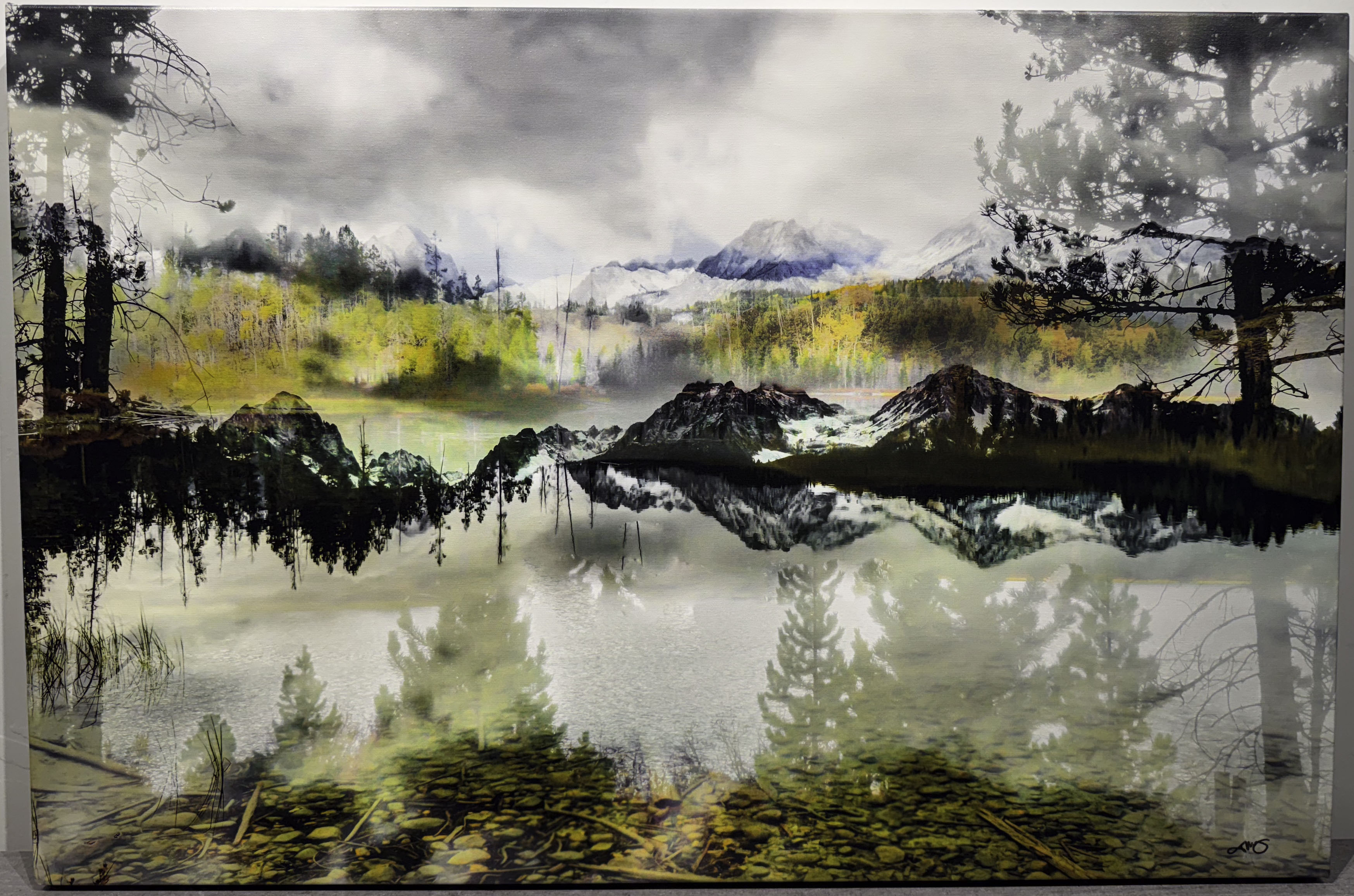 Landscape photograph featuring a reflective mountain lake with forests and distant peaks mirrored in calm water beneath dramatic clouds.