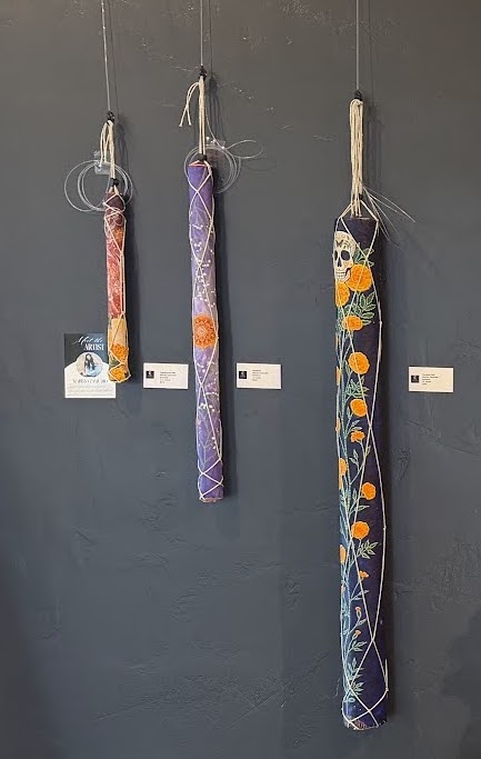 Handcrafted rainsticks by Marissa Zamudio hanging on a dark gallery wall, featuring colorful painted designs. Handcrafted rainsticks by Marissa Zamudio hanging on a dark gallery wall, featuring colorful painted designs.