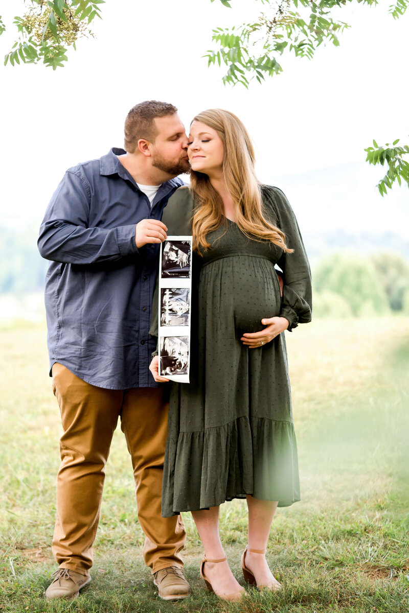 An expecting couple in green and blue kiss while holding their sonogram under a tree on a hill.