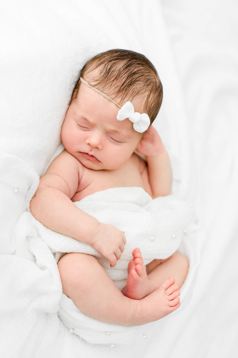 A newborn baby girl sleeps on a white blanket with a matching bow headband.