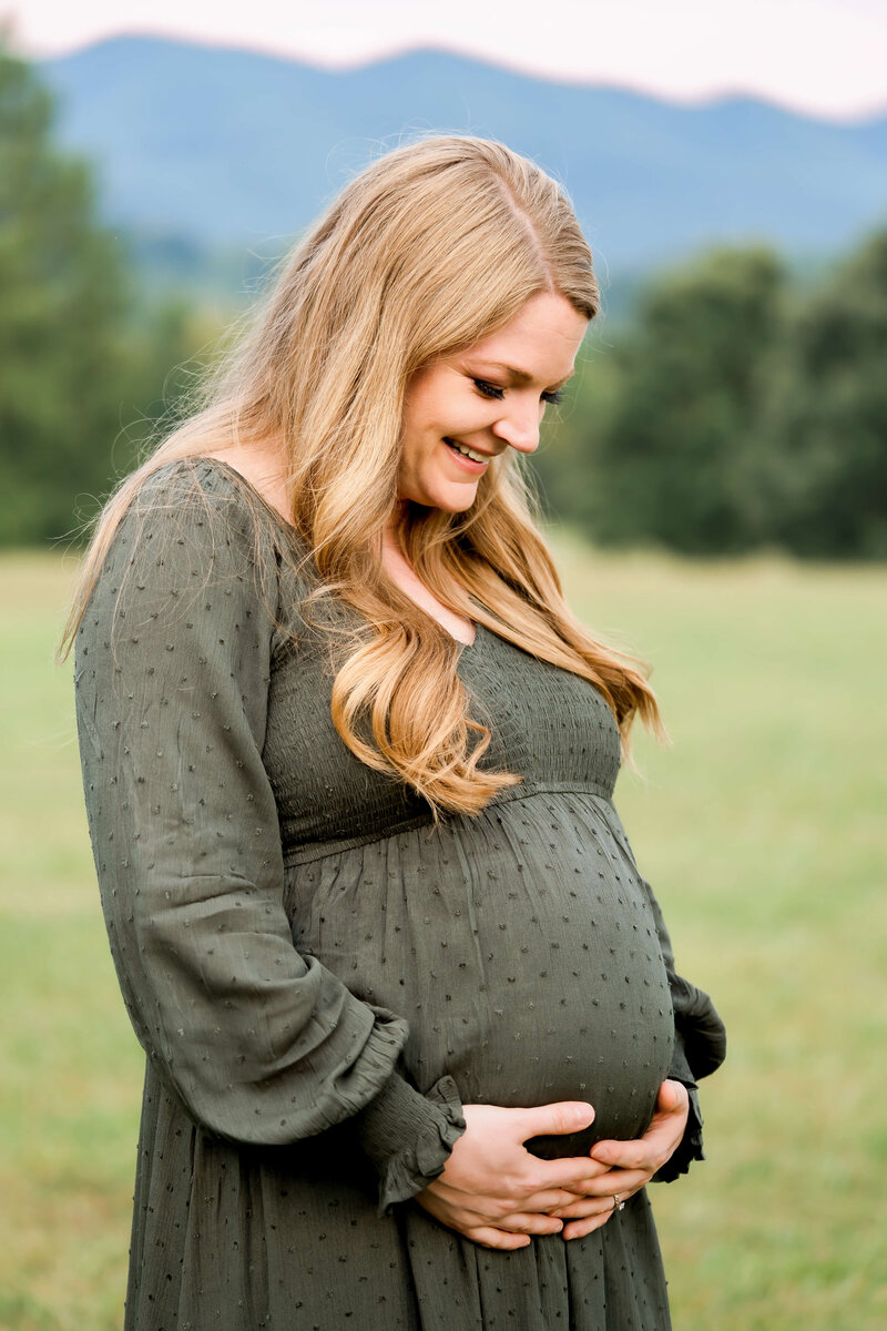 A smiling mom to be in a green maternity gown smiles down to her bump while standing in a high country lawn.