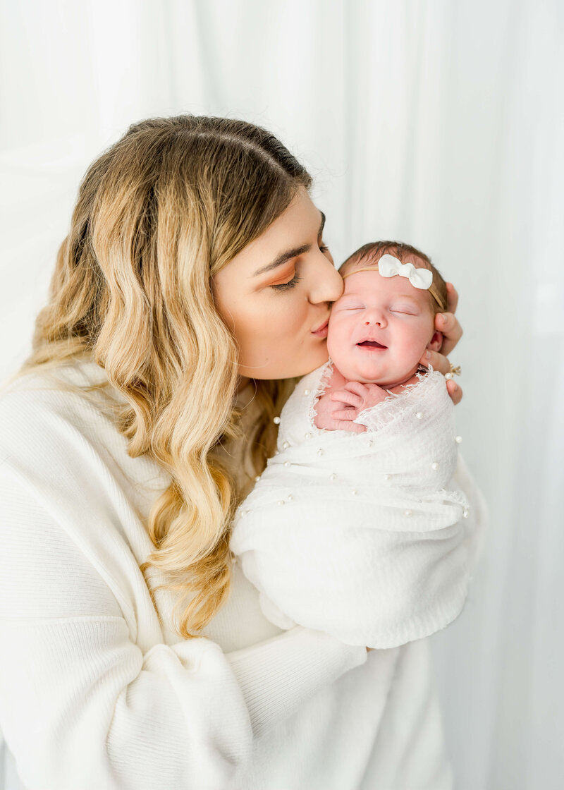 An image from an East Tennessee family photography session of a blonde mom kisses her smiling newborn in her hands in a tight white swaddle.