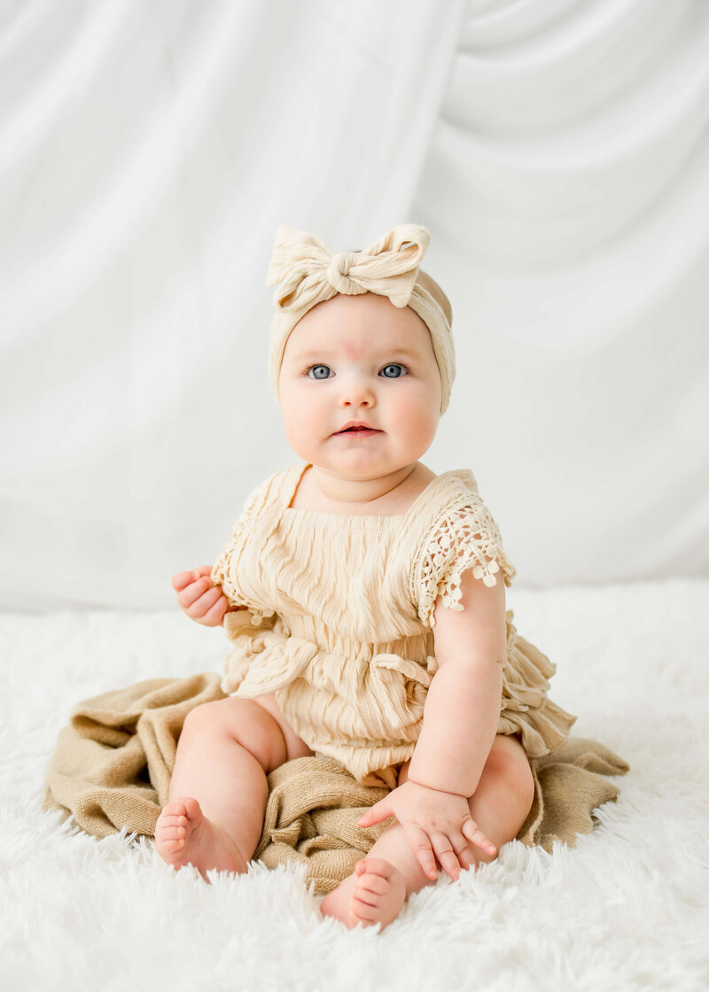A baby girl sits in a studio in a cream lace dress and matching headband.