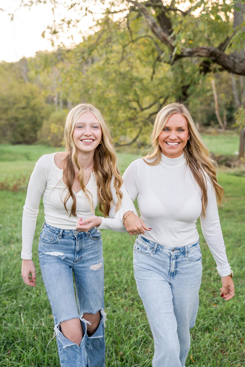 A moment from an East Tennessee family photography session of a mom and daughter in matching white blouse and jeans walking with arms linked on the edge of a forest with big smiles.