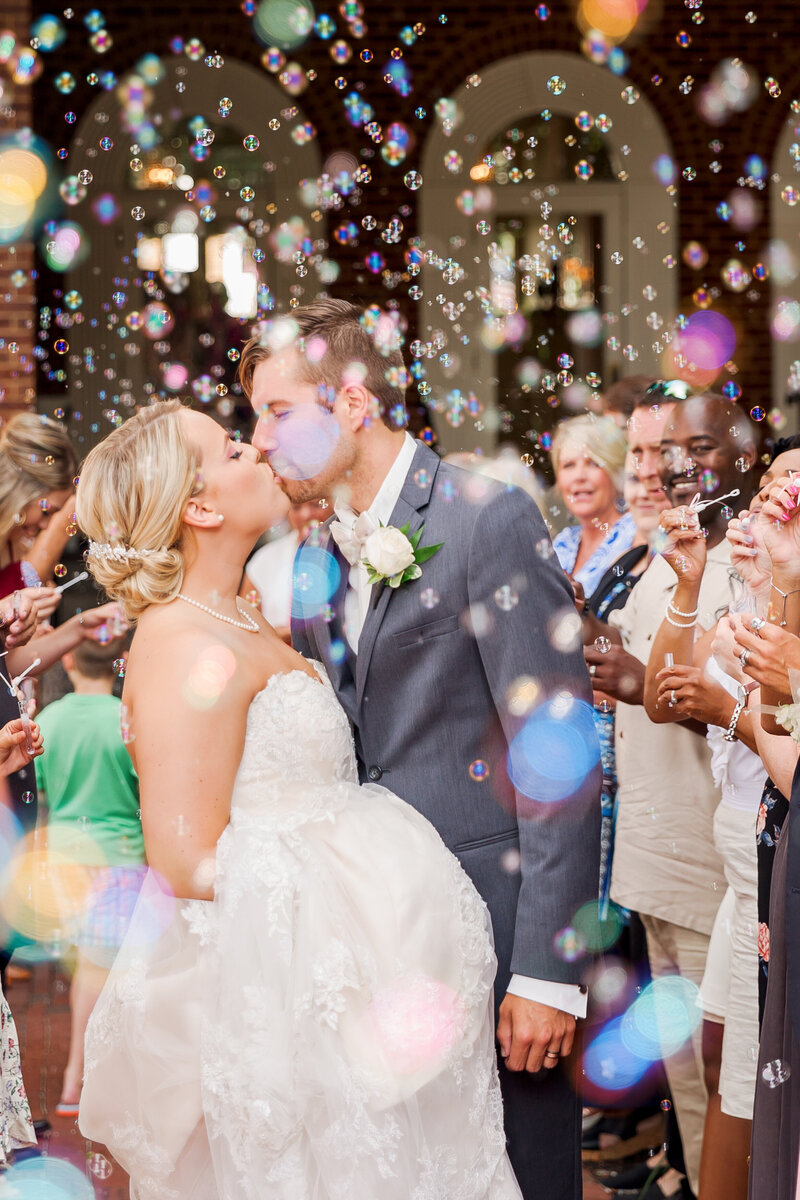 A bride and groom kiss while guests blow bubbles all around them during their exit.