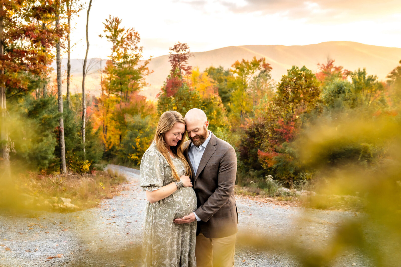 Expecting parents smile while cradling the bump in a green dress and brown jacket at sunset in the fall.
