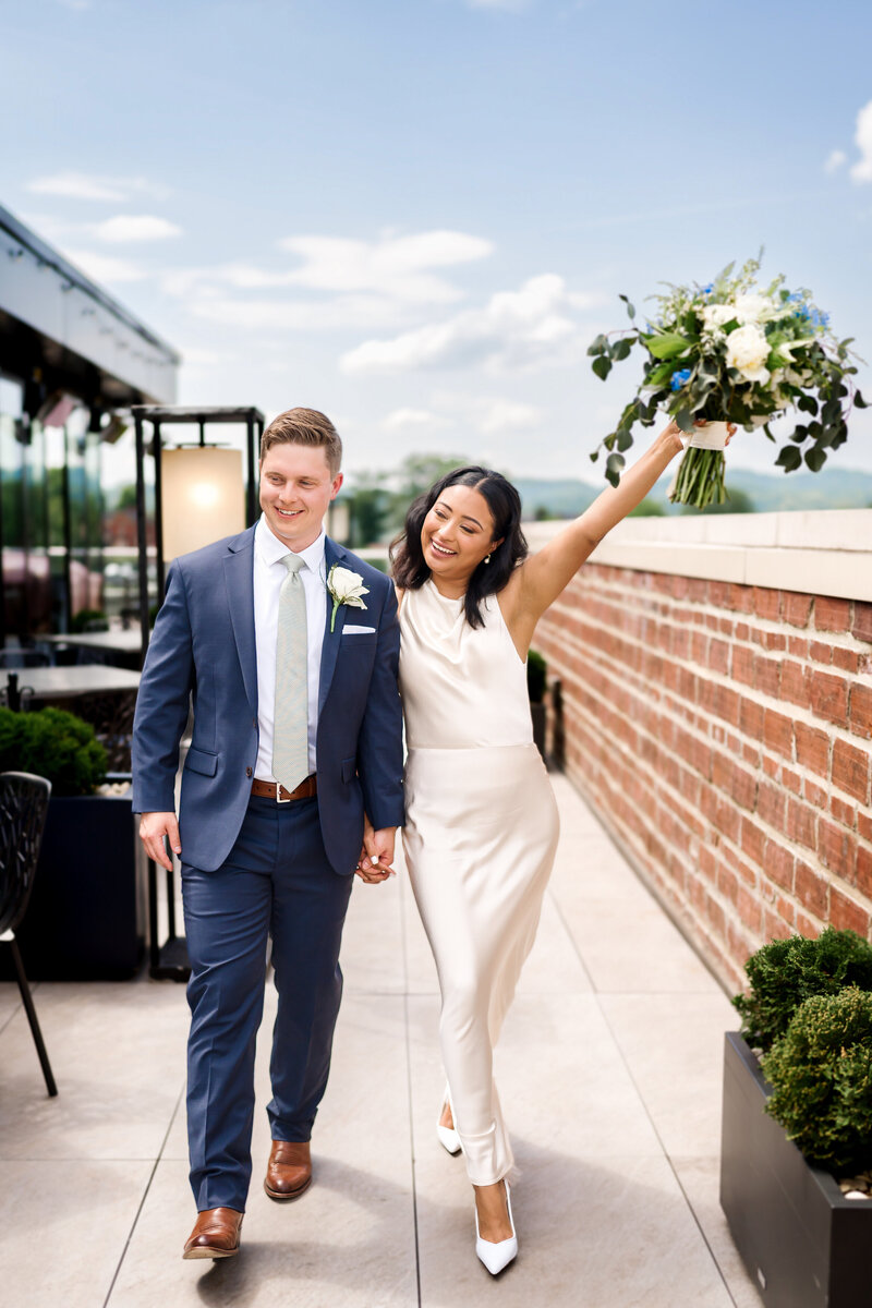 A moment from an East Tennessee wedding photography gallery of a bride lifting her bouquet above her head while walking on a patio holding hands with her groom in blue.
