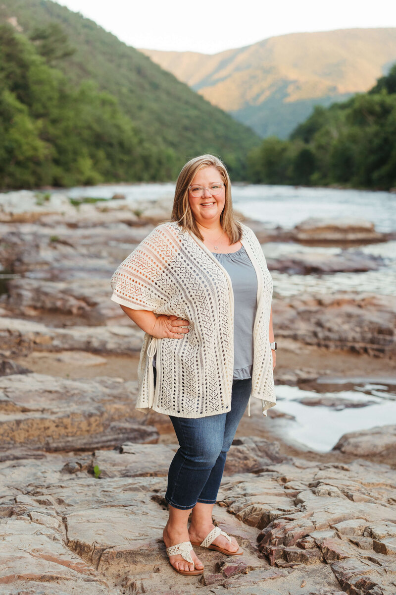 East Tennessee family photographer April Booher stands with a hand on her hip in jeans and white knit sweater on the edge of a river at sunset.