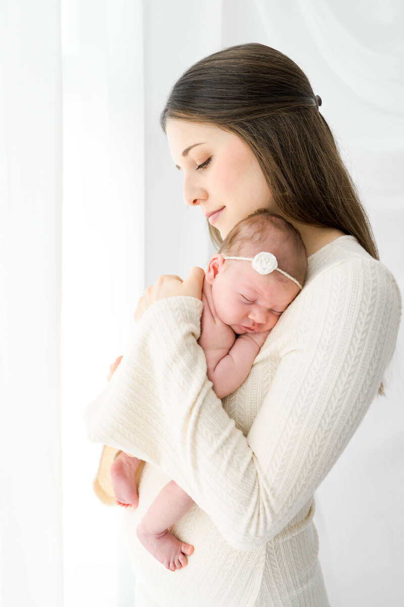 A moment from an East Tennessee newborn photographer of a newborn baby sleeping against mom's shoulder in a tiny flower headband while mom stands in a window.