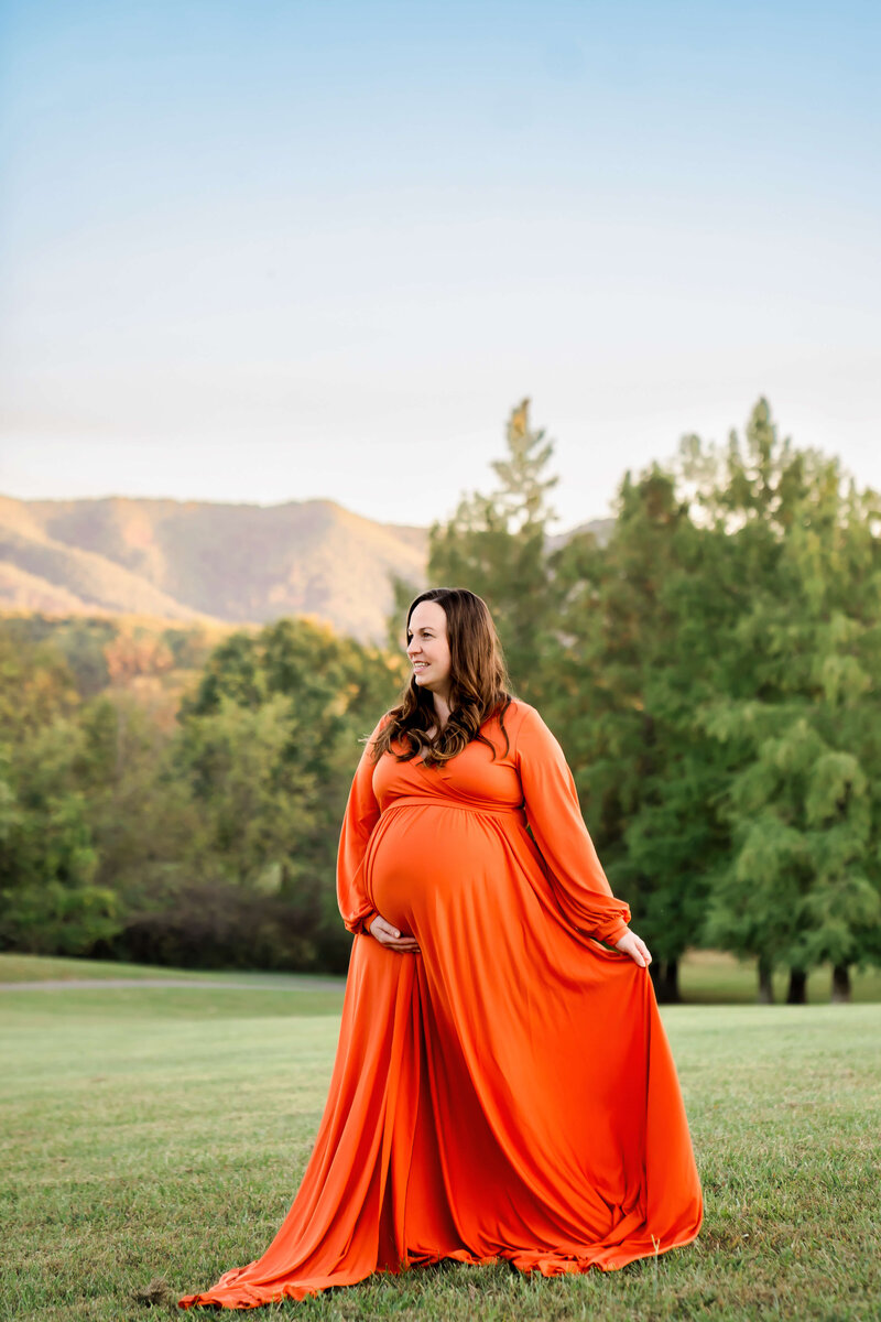 An East Tennessee family photographer captured moment of a mom to be walking with a hand under her bump in a mountain lawn in a red maternity gown.