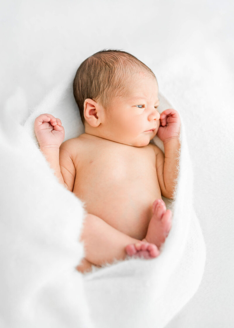 A newborn baby lays naked in a white blanket with eyes open.