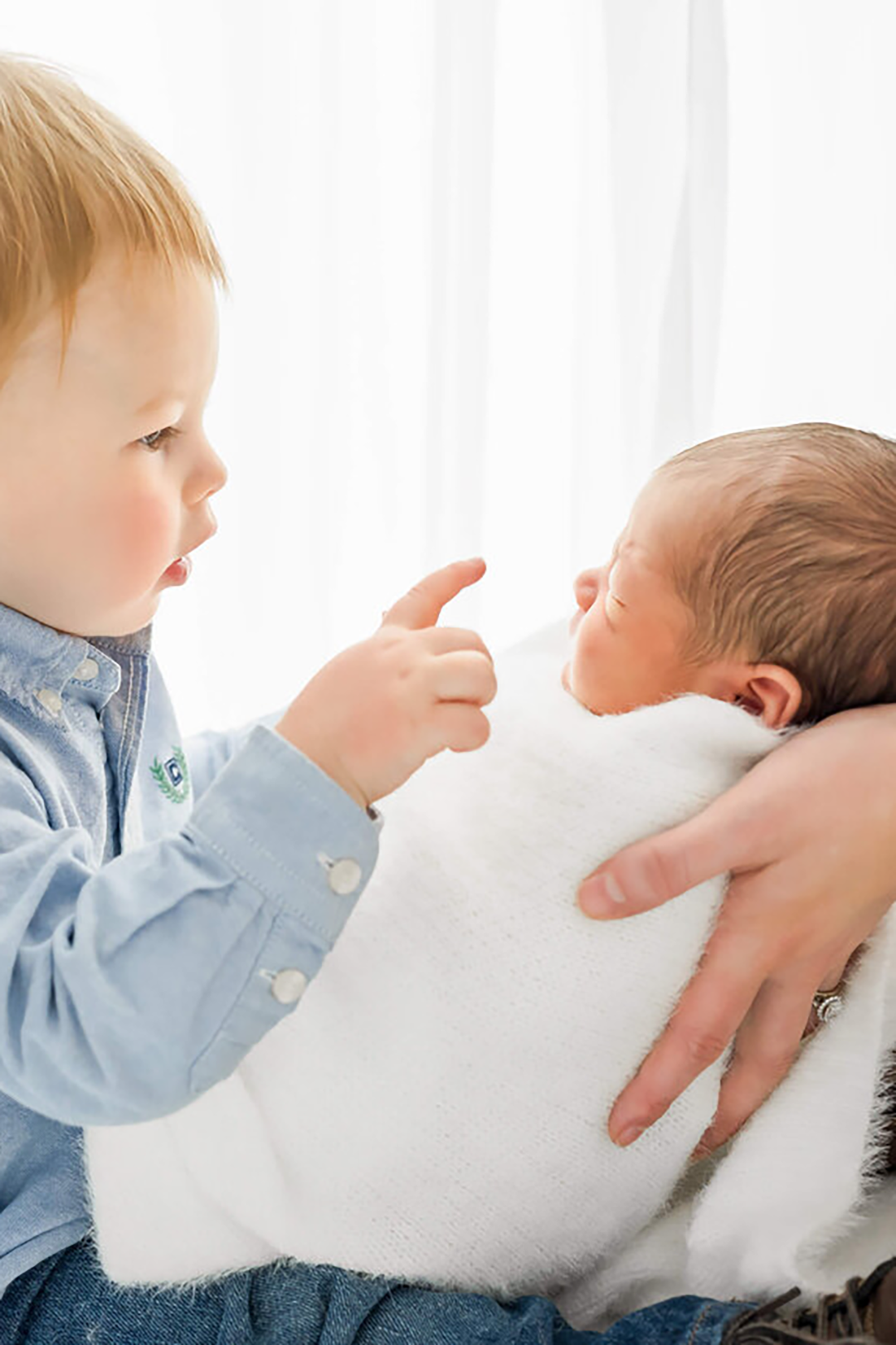 A toddler boy reaches to touch his newborn baby sibling being help in his lap by mom in a white swaddle.