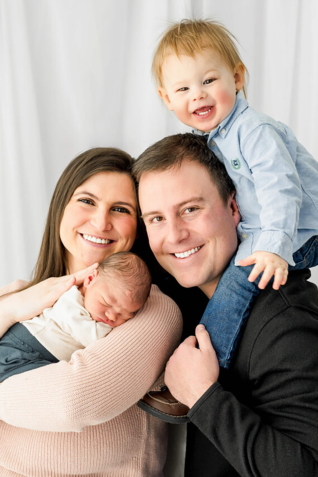 A smiling mom and dad sit in a studio with a giggling toddler on dad's shoulders and sleeping newborn on mom's.