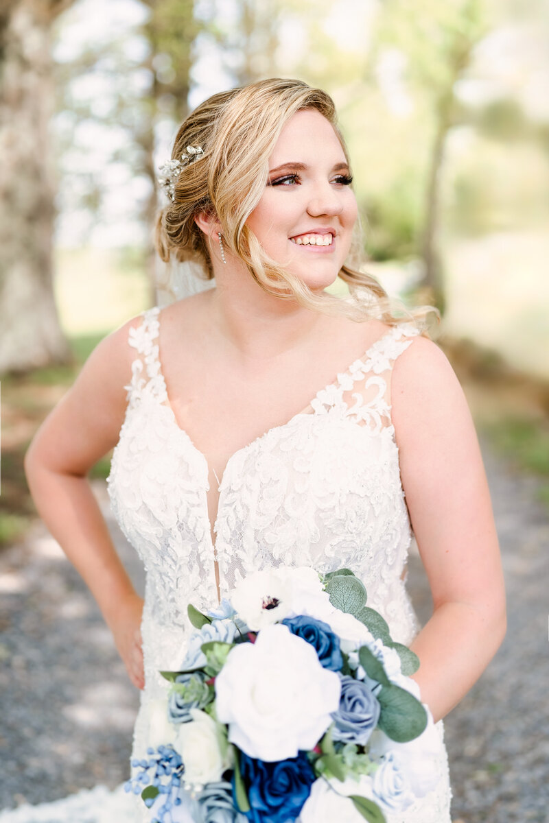 A happy bride smiles over her shoulder while standing in a forest holding her white and blue bouquet