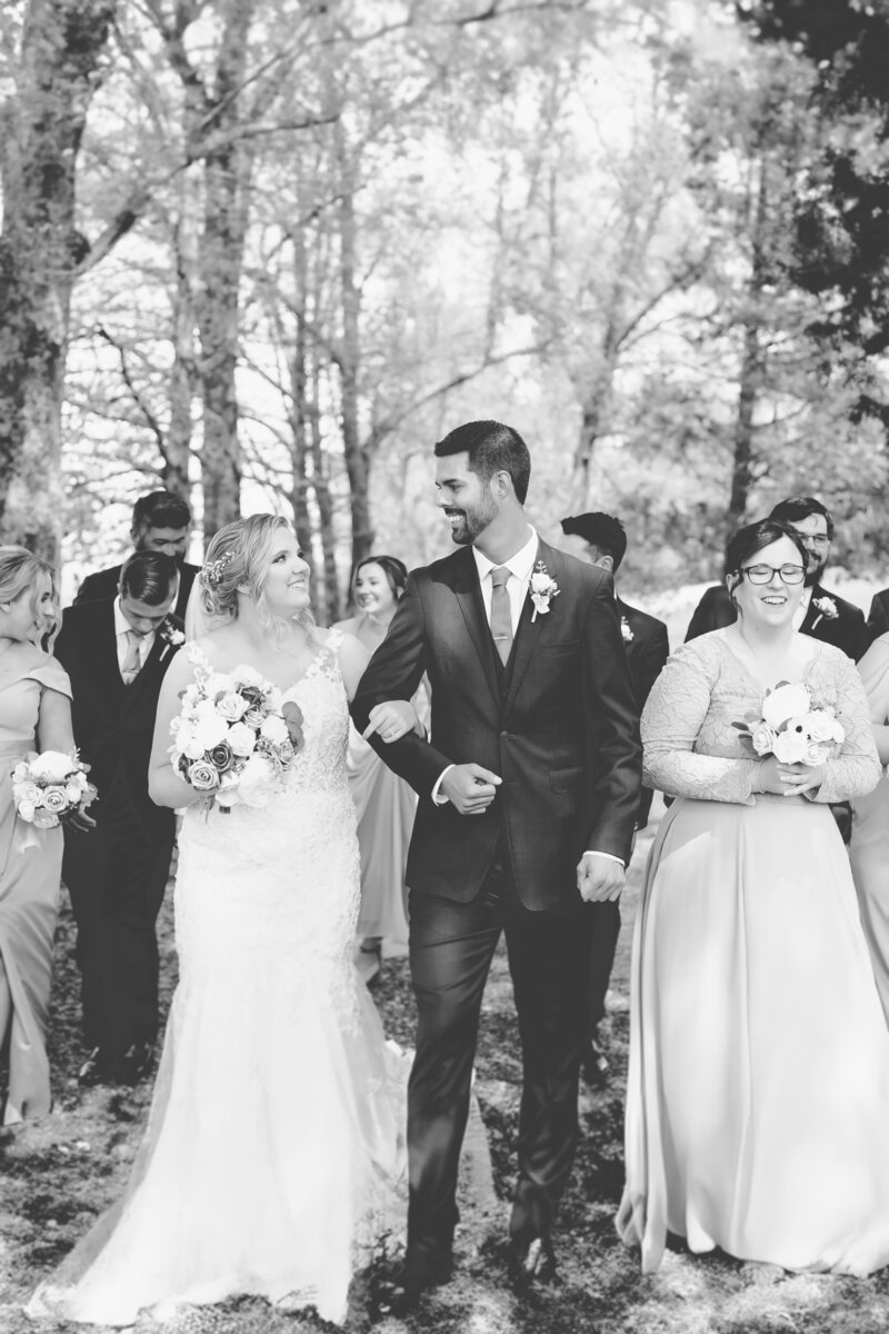 A bride and groom walk in a forest with their wedding party in black and white.