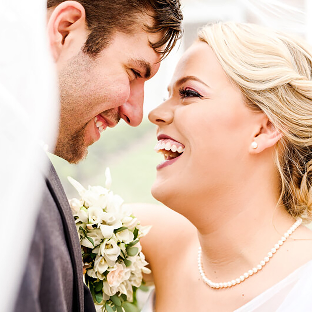 A bride and groom laugh and smile while hiding under the veil.