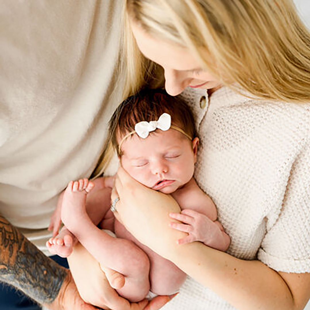 New mom and dad snuggle while on a couch cradling their sleeping newborn girl wearing a white bow headband.