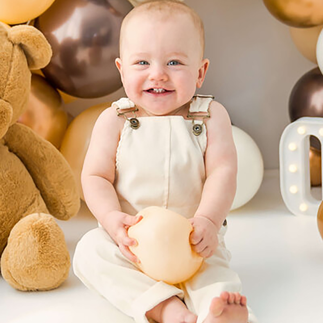 A little baby smiles while holding. balloon while sitting next to a stuffed teddy bear. 