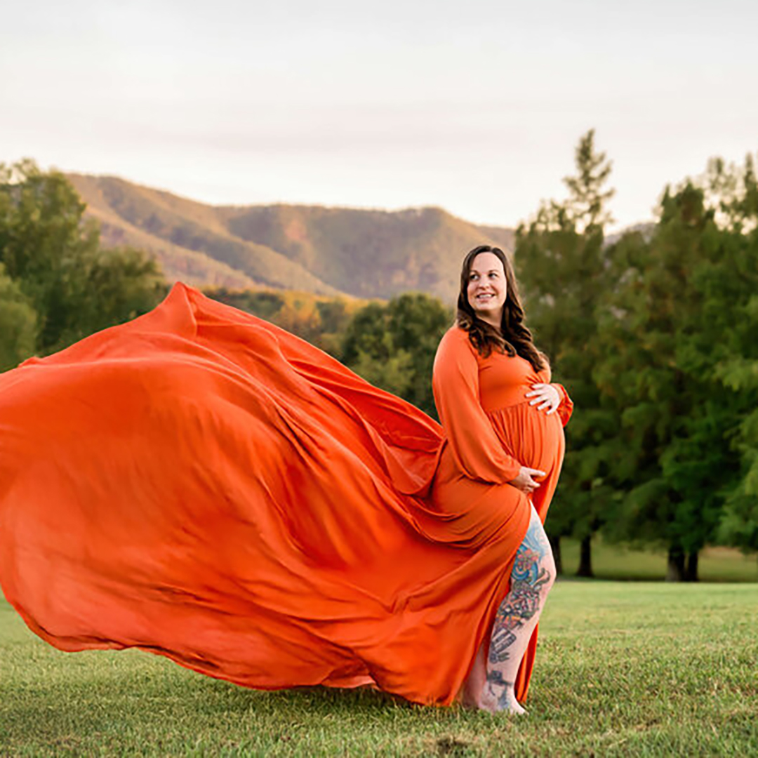 A mother to be in a long flowing maternity gown stands in a field at sunset with hands on her bump.