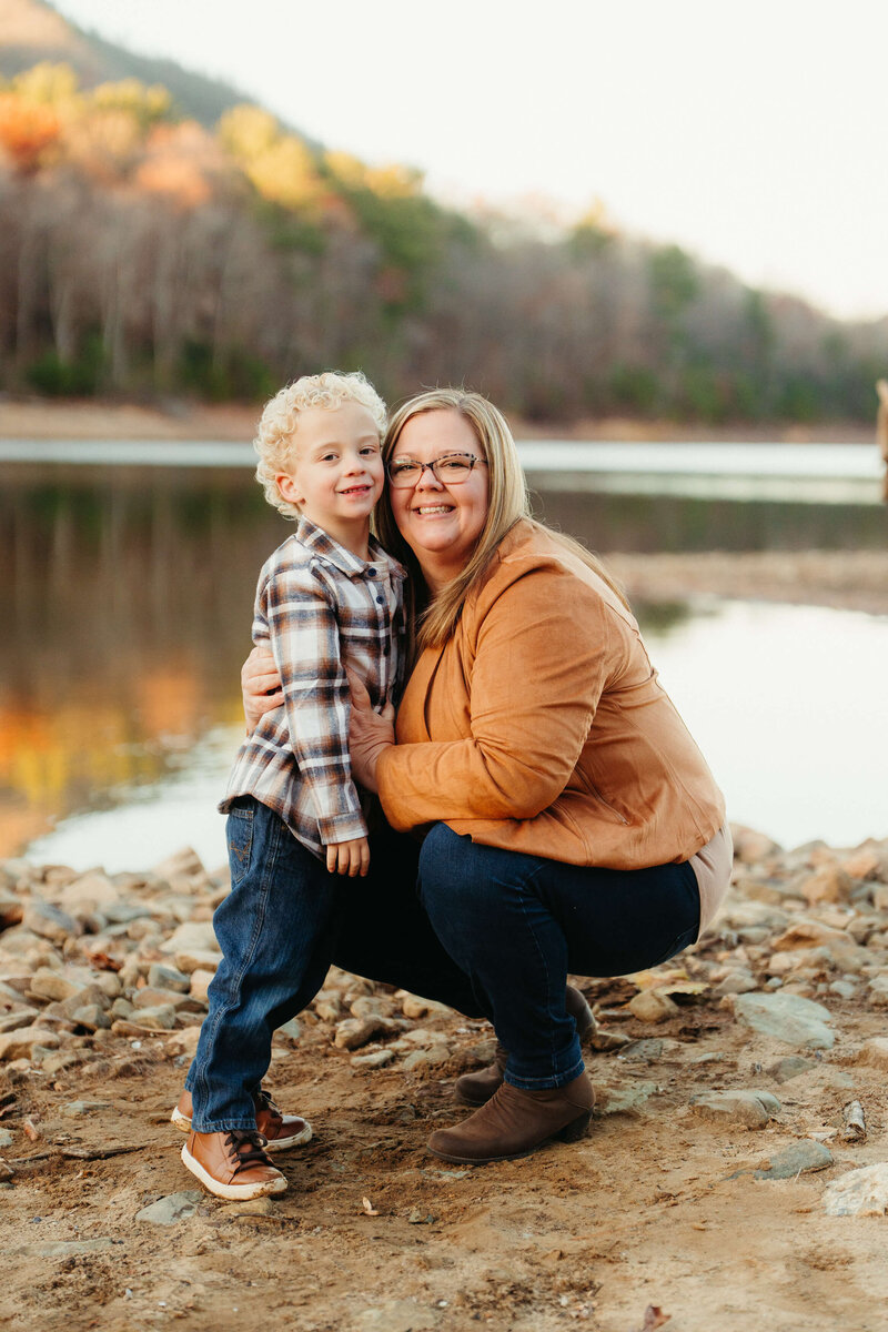 April Booher hugs her toddler son in a plaid shirt and jeans on the edge of a lake in fall at sunset.