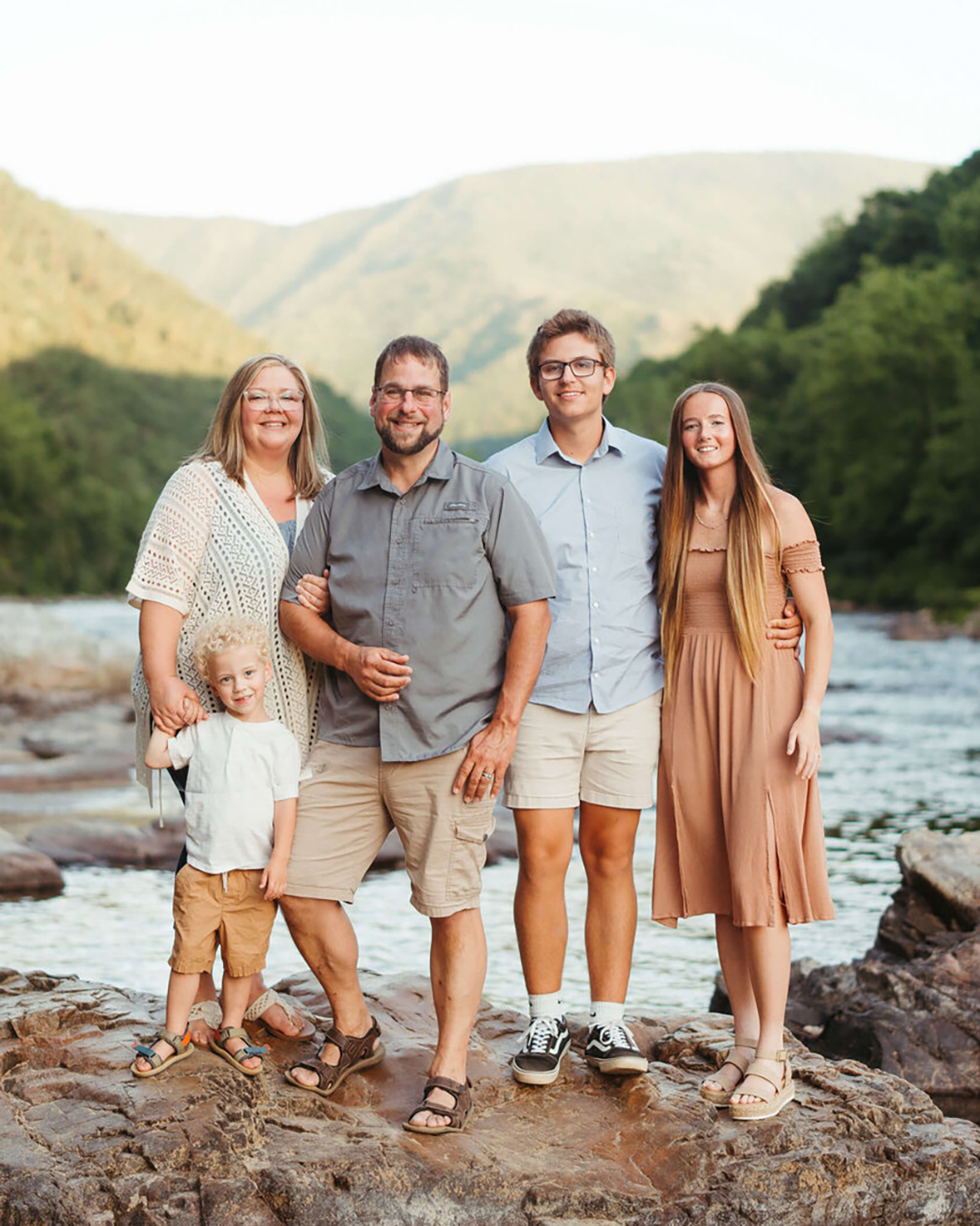 A mom and dad stand on a boulder in natural tone outfits with their two teen children and toddler son at sunset in the mountains.